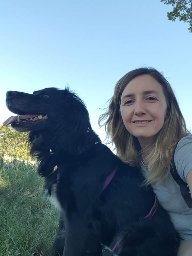 Femme souriante et chien noir avec harnais violet, langue pendante, posant en selfie sous un ciel bleu clair.