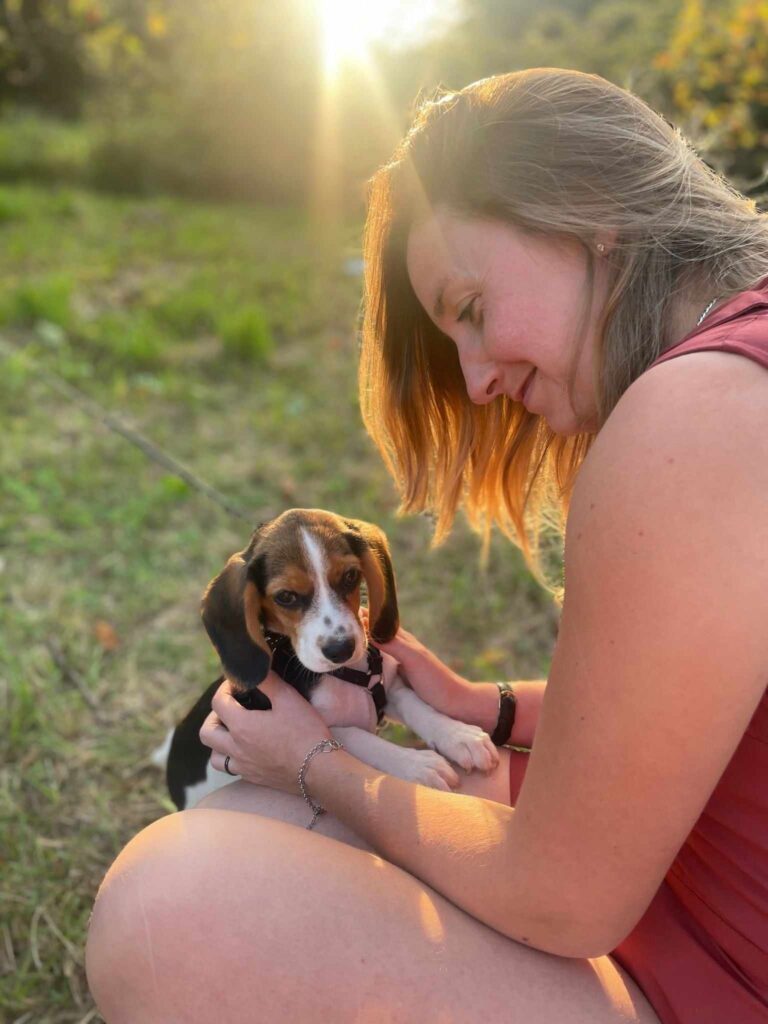 Une femme souriante et aux cheveux dorés caresse un adorable chiot beagle tricolore, regardant le spectateur, dans un champ ensoleillé.