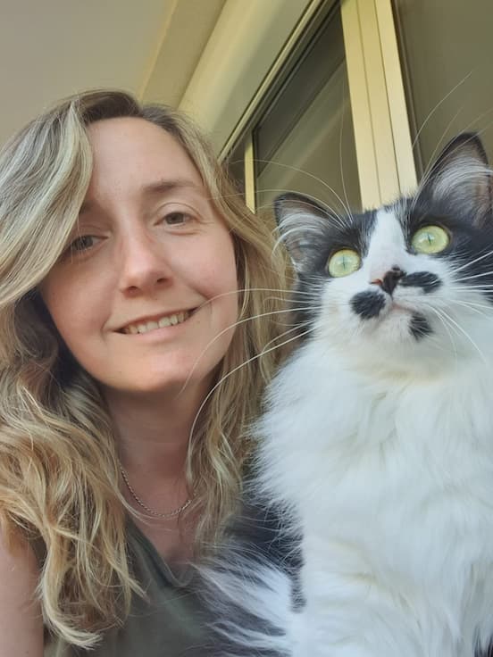 Femme souriante aux cheveux blonds posant avec un chat noir et blanc aux yeux verts. Le chat a une tache noire distinctive sous le nez.